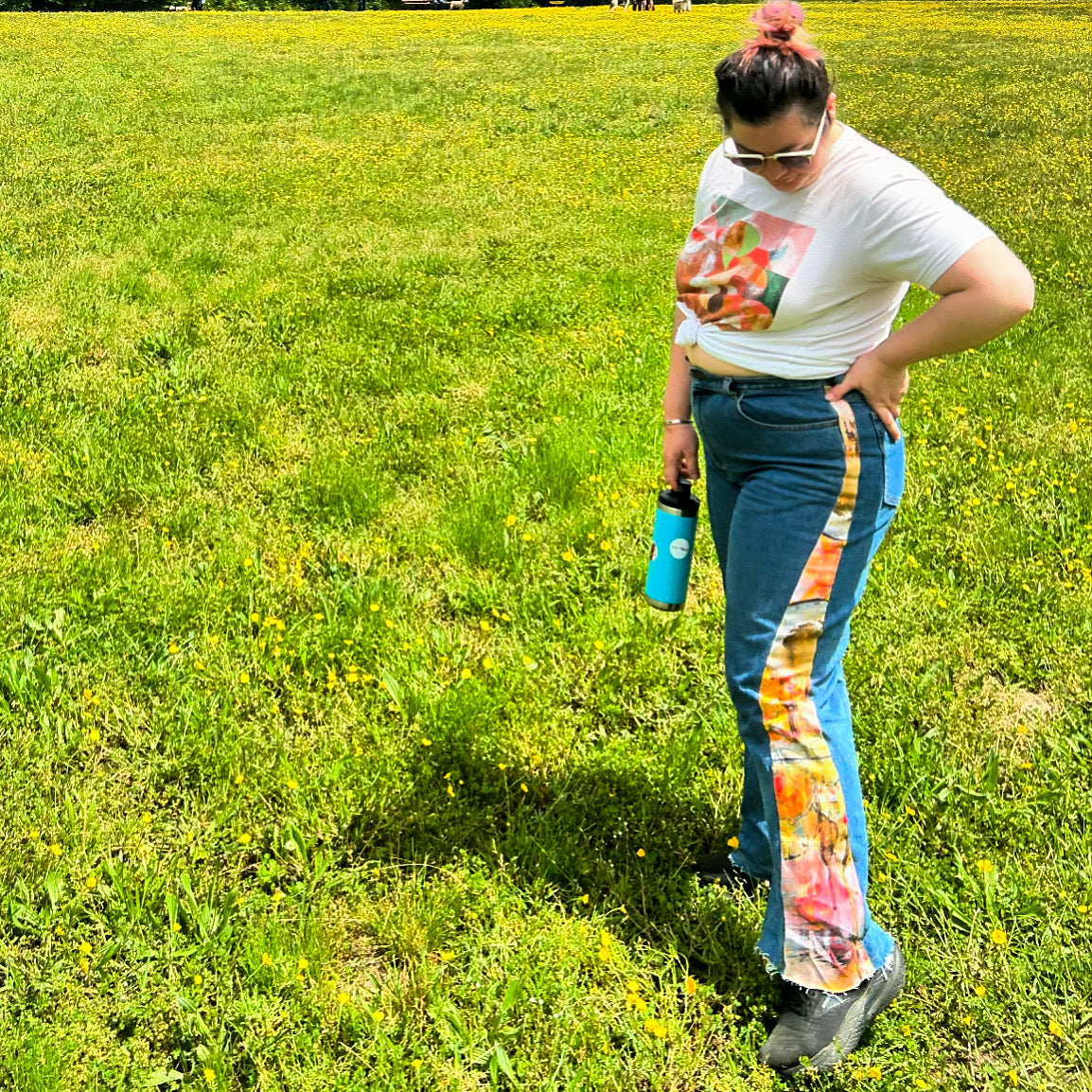A person standing on grass wearing a white t-shirt with a colorful floral design on the top, flared jeans with a colorful floral design on the side panels, and sunglasses.