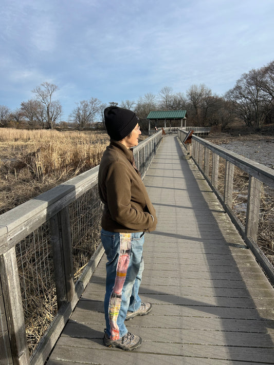A person standing on a boardwalk wearing a beanie, a jacket, and jeans with a unique design on the side panels.