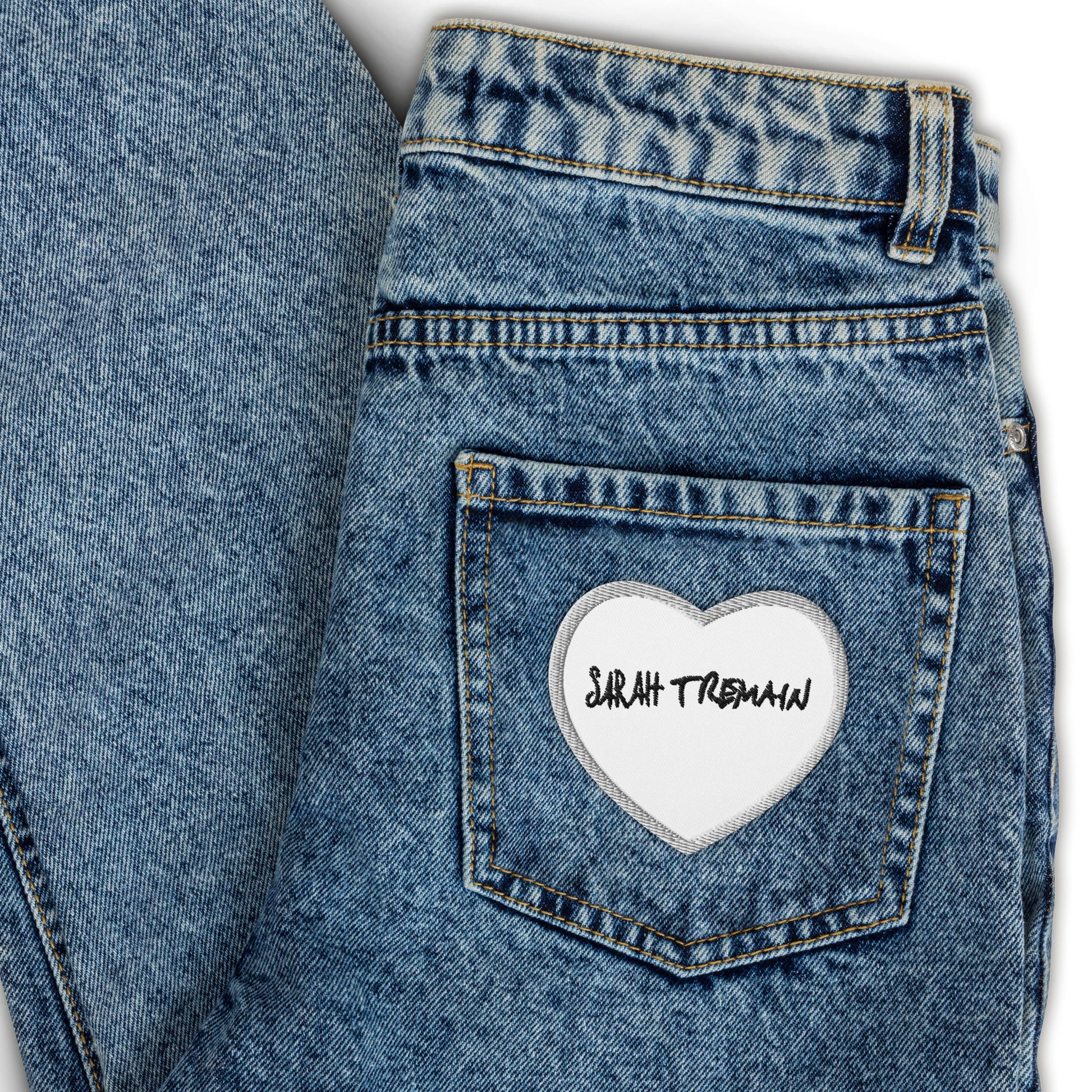 A white iron-on patch with the text 'SARAH TREMAIN' inside a heart-shaped design, placed on the back pocket of blue denim jeans.