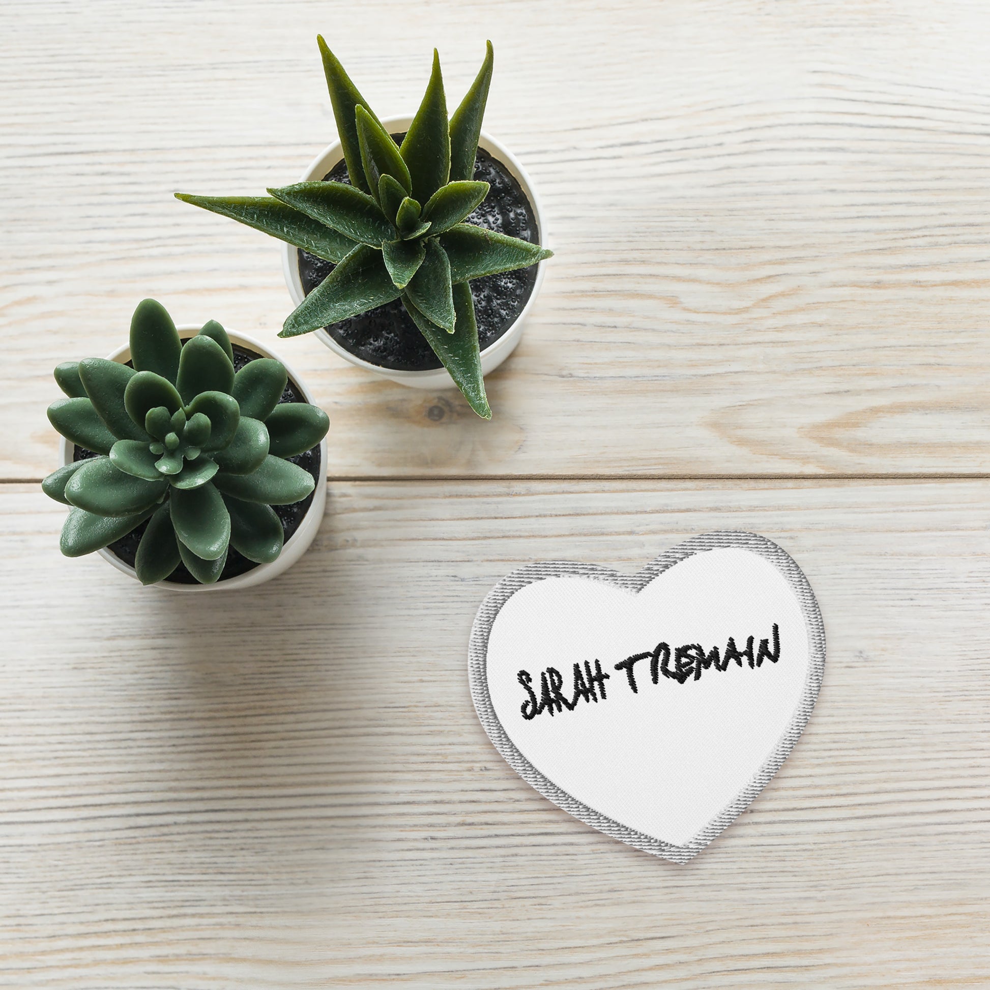 A white iron-on patch with the text 'SARAH TREMAIN' inside a heart-shaped design, placed on table next to plants