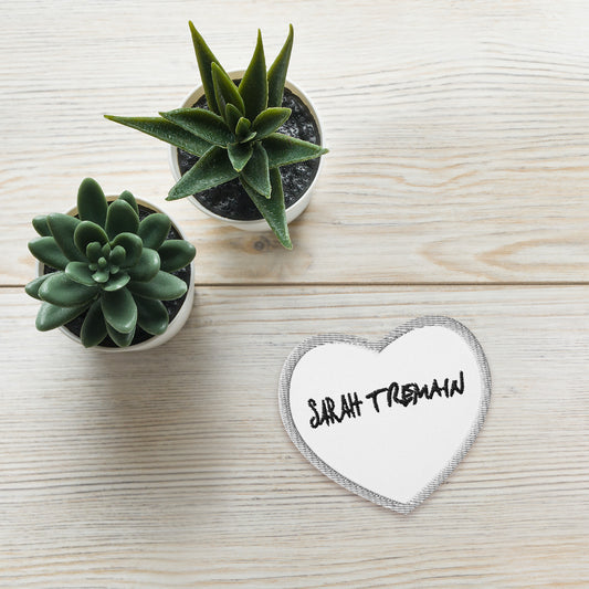 A white iron-on patch with the text 'SARAH TREMAIN' inside a heart-shaped design, placed on table next to plants