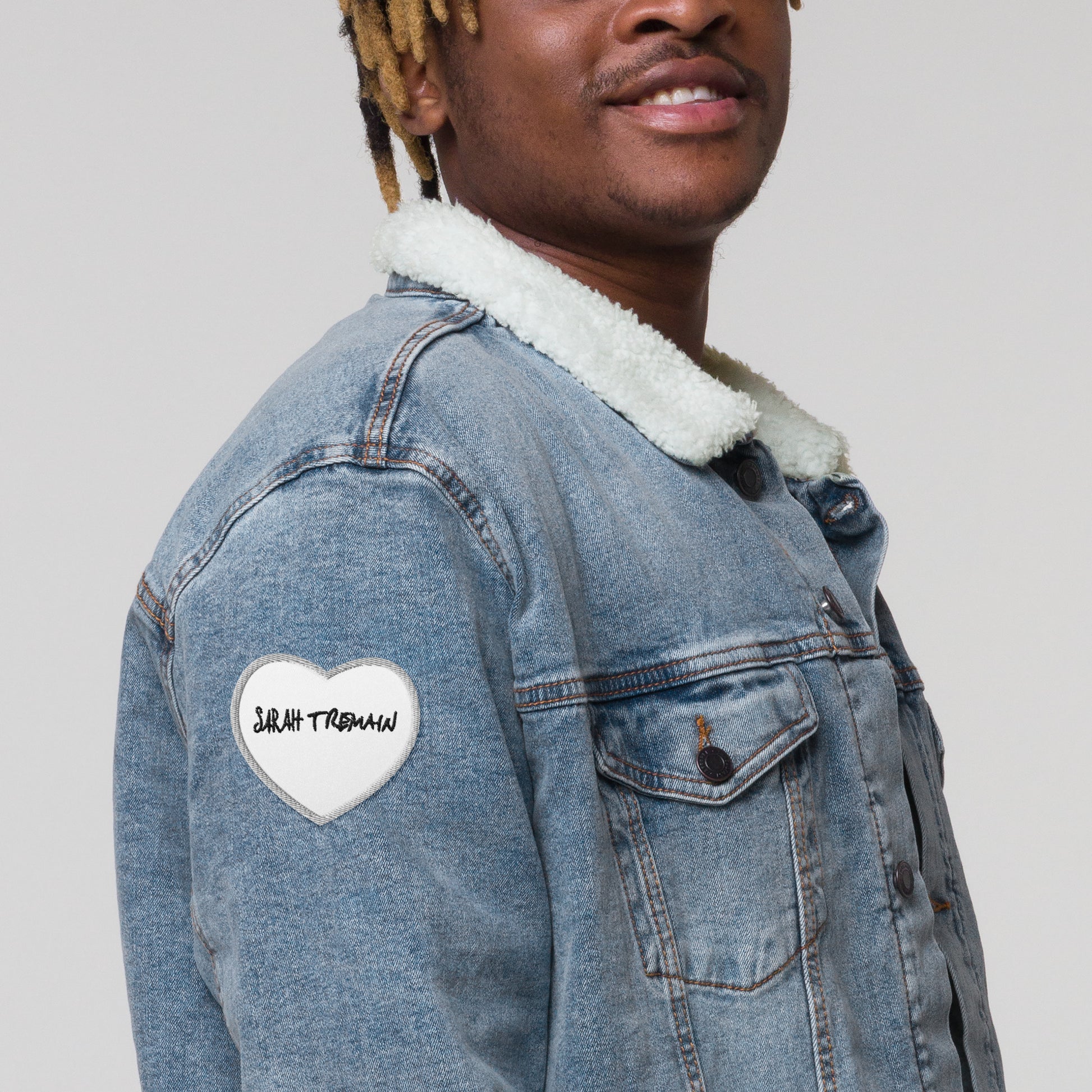 A white iron-on patch with the text 'SARAH TREMAIN' inside a heart-shaped design, shown on denim jacket on model