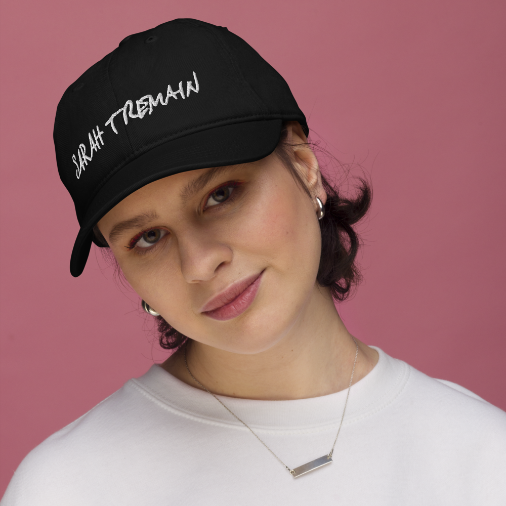 A person wearing a black organic dad hat with the name 'SARAH TREMAIN' embroidered on the front.
