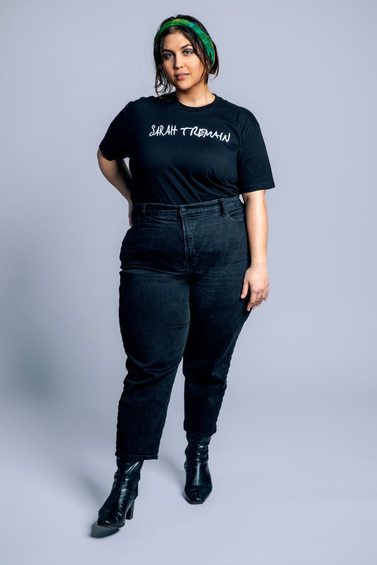 A woman wearing a black t-shirt with the text 'SARAH TREMAIN' on the front and blue jeans, paired with black boots.
